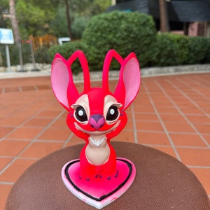 Może przedstawiać: R&oacute;żowa i czerwona figurka Stitch stojąca na r&oacute;żowej podstawie w kształcie serca. Figurka ma duże oczy i biały kołnierzyk.