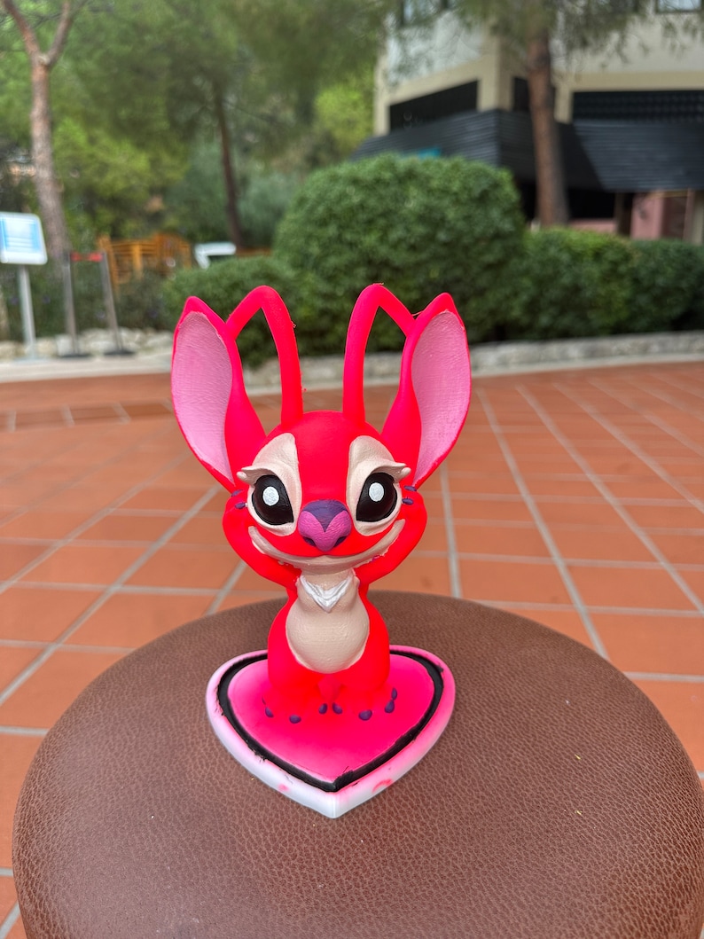 Może przedstawiać: R&oacute;żowa i czerwona figurka Stitch stojąca na r&oacute;żowej podstawie w kształcie serca. Figurka ma duże oczy i biały kołnierzyk.