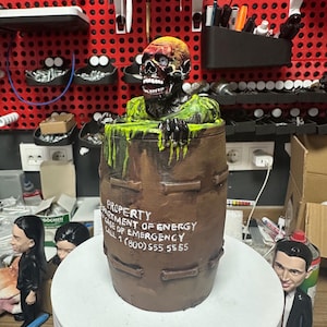 Pode incluir: Um barril castanho com um zumbi emergindo de uma gosma verde. O zumbi tem um rosto em decomposição com dentes expostos e uma cabeça amarela e vermelha. O barril tem o texto "PROPERTY DEPARTMENT OF ENERGY CASE OF EMERGENCY CALL 1 (800) 555 5555".