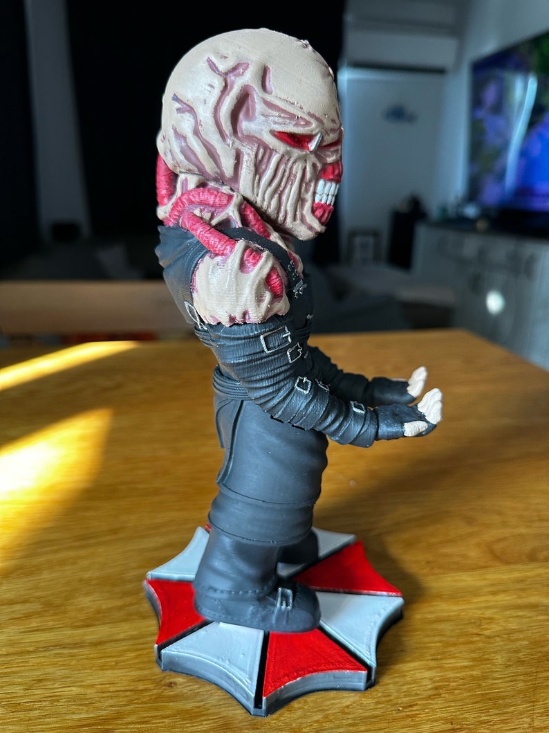 Pode incluir: Uma figura detalhada de um personagem da s&eacute;rie de videogames Resident Evil. A figura est&aacute; em p&eacute; sobre uma base com a forma do logotipo da Umbrella Corporation. O personagem est&aacute; vestindo uma roupa preta e tem uma cabe&ccedil;a grotesca e carnuda com olhos vermelhos.
