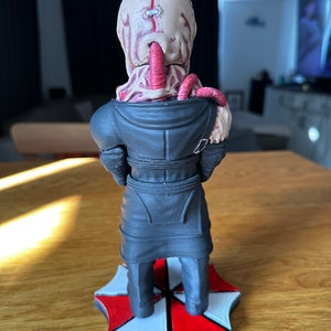 Pode incluir: Uma figura impressa em 3D de um personagem da s&eacute;rie de videogames Resident Evil. O personagem est&aacute; vestindo um casaco preto e tem uma base de guarda-chuva vermelha e branca.