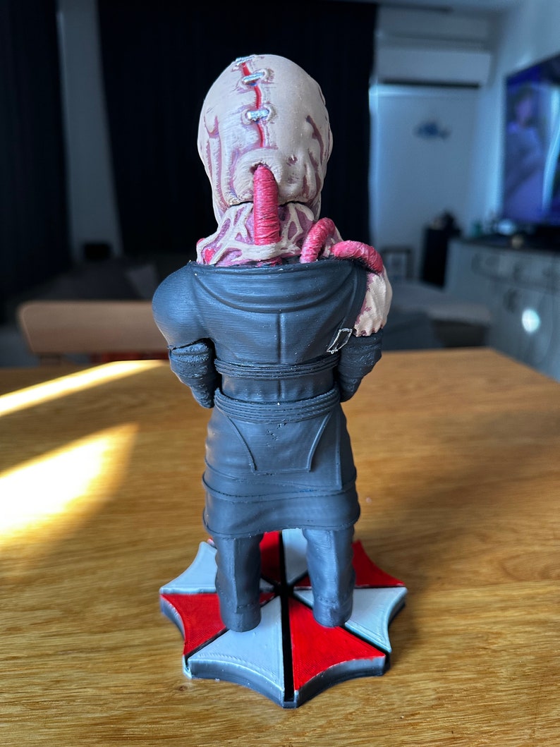 Pode incluir: Uma figura impressa em 3D de um personagem da s&eacute;rie de videogames Resident Evil. O personagem est&aacute; vestindo um casaco preto e tem uma base de guarda-chuva vermelha e branca.