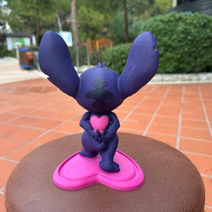 May include: A blue 3D printed figurine of Stitch from Lilo & Stitch holding a pink heart. The figurine is standing on a pink heart-shaped base.