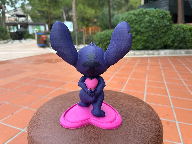 May include: A blue 3D printed figurine of Stitch from Lilo & Stitch holding a pink heart. The figurine is standing on a pink heart-shaped base.