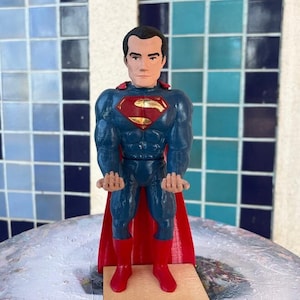 May include: A blue and red Superman action figure with a gold emblem on the chest. The figure is standing on a wooden base.