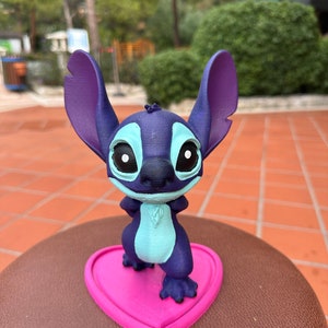 May include: A blue and purple Stitch figurine standing on a pink heart-shaped base. The figurine has large ears, big eyes, and a mischievous expression.