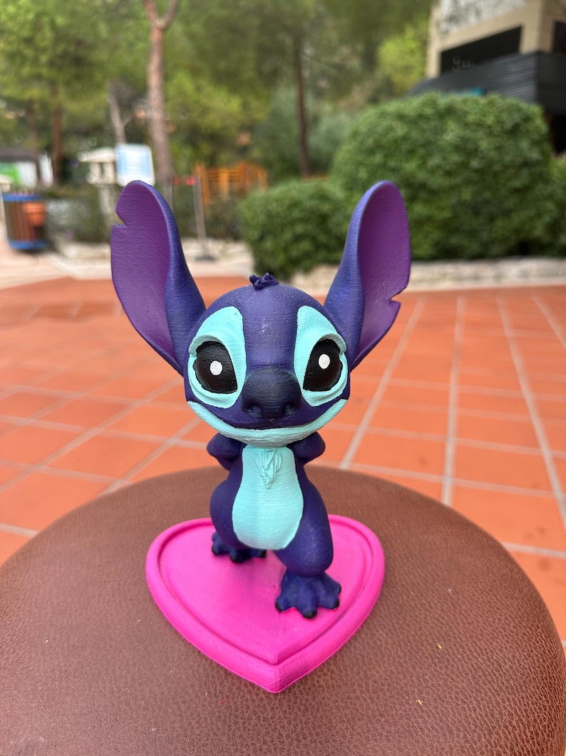 May include: A blue and purple Stitch figurine standing on a pink heart-shaped base. The figurine has large ears, big eyes, and a mischievous expression.