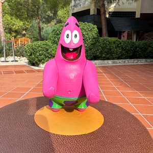 Puede incluir: Un personaje de dibujos animados rosa con una gran sonrisa y ojos grandes. El personaje lleva pantalones cortos verdes con lunares morados y está de pie sobre un círculo amarillo.