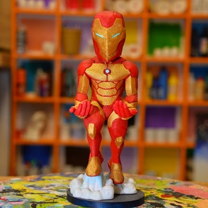Puede incluir: Una figura de Iron Man roja y dorada con una base de nube azul y blanca. La figura está de pie con los brazos extendidos.
