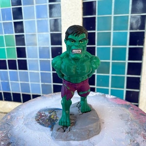 Könnte beinhalten: Eine grüne Hulk-Figur mit einem lila-weißen Lendenschurz, die auf einem grauen Felssockel steht. Die Figur hat ein detailliertes Gesicht mit einem wütenden Ausdruck.