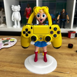 Puede incluir: Un controlador amarillo de PlayStation 4 está sostenido por una figura de un personaje de dibujos animados con cabello largo y rubio, un vestido azul y blanco y botas rojas. La figura está de pie sobre una base blanca.