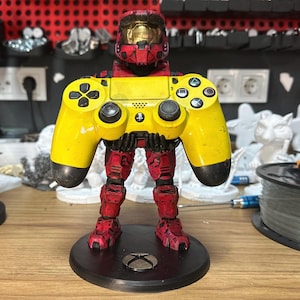 May include: A yellow and black video game controller is held by a red and black action figure of a soldier wearing a helmet. The figure is standing on a black circular base.
