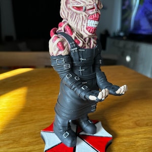 Pode incluir: Um suporte de telefone tem&aacute;tico Resident Evil preto e vermelho com um personagem com um rosto grotesco e carnudo e um traje de couro preto. O personagem est&aacute; em p&eacute; sobre um logotipo de guarda-chuva vermelho e branco.