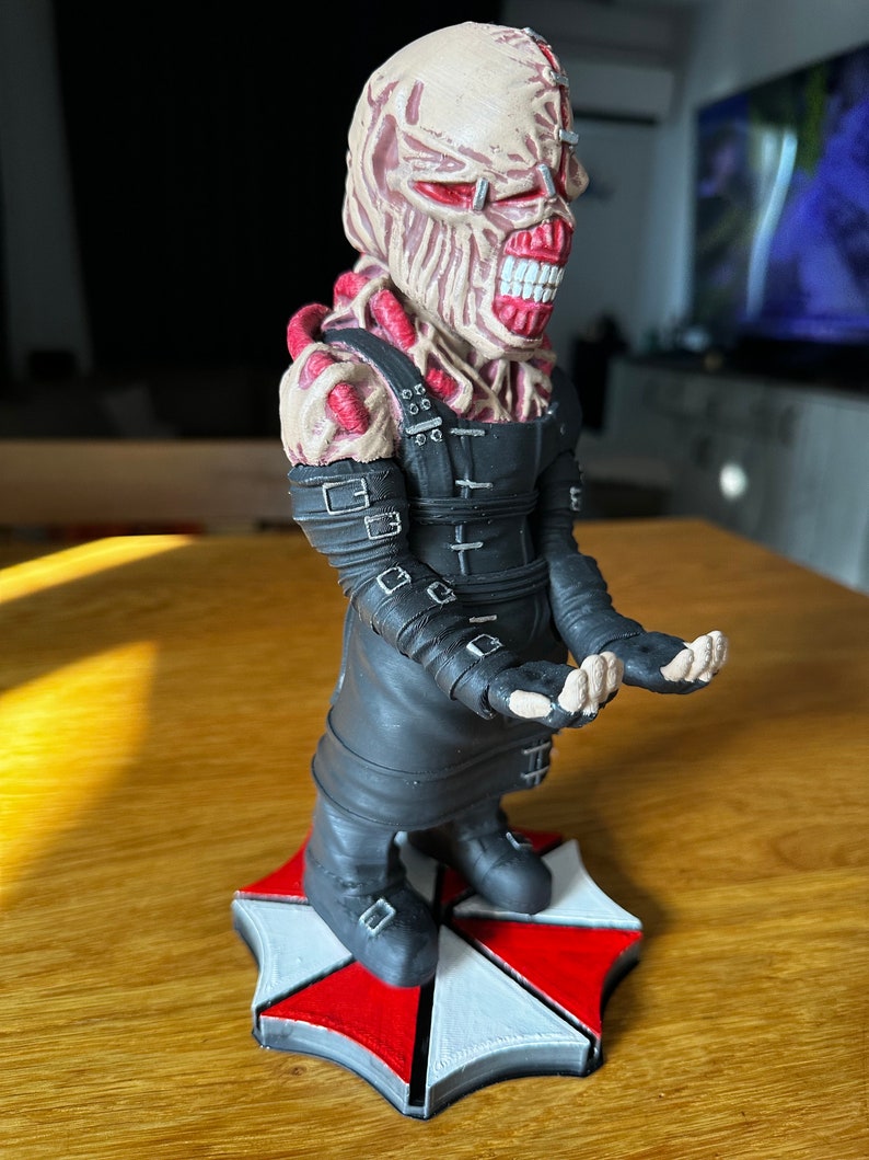 Pode incluir: Um suporte de telefone tem&aacute;tico Resident Evil preto e vermelho com um personagem com um rosto grotesco e carnudo e um traje de couro preto. O personagem est&aacute; em p&eacute; sobre um logotipo de guarda-chuva vermelho e branco.