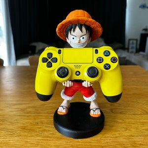 May include: A yellow PlayStation 4 controller is held by a figurine of a cartoon character wearing a red and white outfit and an orange hat. The figurine is standing on a black base.
