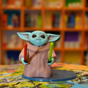 May include: A blue and green 3D printed figurine of a character from the Star Wars franchise, holding two lightsabers. The figurine is standing on a blue circular base.