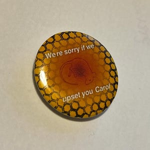 May include: A round button with a honeycomb pattern in yellow and black. The center has a brown rat illustration and the text "We're sorry if we upset you Carol".