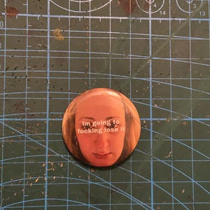 May include: A circular badge featuring a woman's face and the text "im going to fucking lose it" in white. The badge is set against a green cutting mat with grid lines.