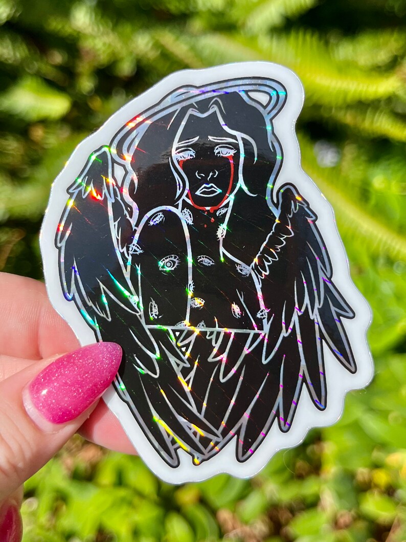 May include: A black and white sticker of an angel with wings, a halo, and eyes all over its body. The angel is crying red tears. The sticker has a holographic finish.