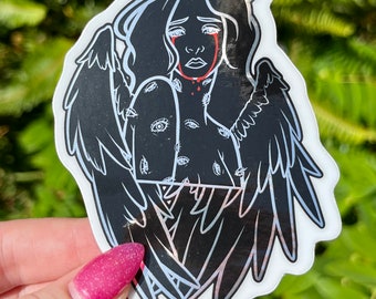 May include: A black sticker featuring a crying angel with a halo and wings. The angel has red tears and multiple eyes on its torso. The sticker has a white border and is held against a green background.