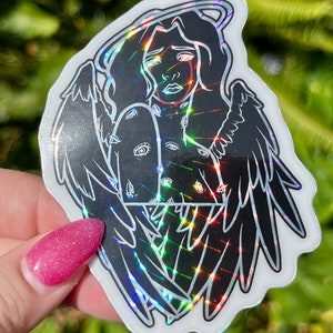 May include: A black and white sticker of an angel with wings, a halo, and eyes covering the body. The sticker has a holographic finish.
