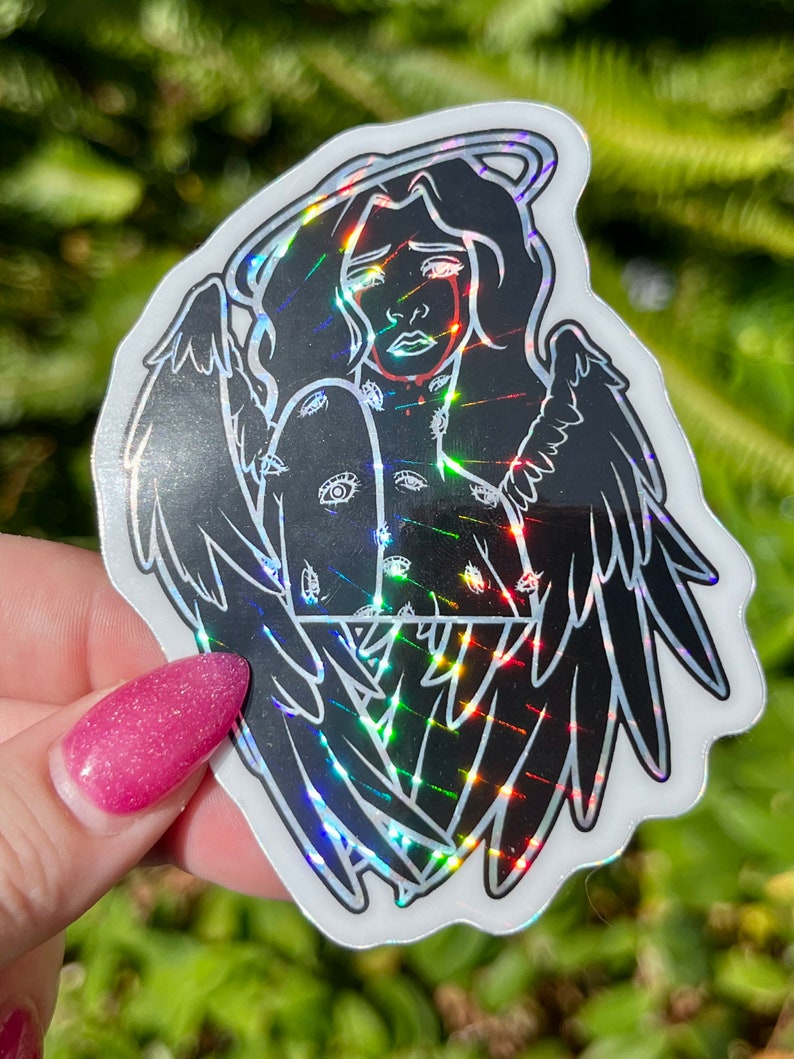 May include: A black and white sticker of an angel with wings, a halo, and eyes covering the body. The sticker has a holographic finish.