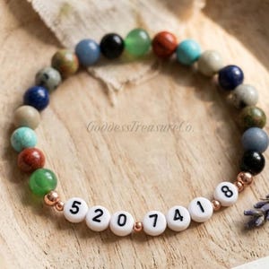 May include: A colourful gemstone bead bracelet. The bracelet features white letter beads spelling out the numbers "5207418". The bracelet is presented on a wooden surface.