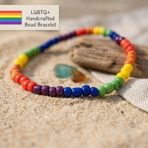 Może przedstawiać: Ręcznie robiona bransoletka z koralików LGBTQ+. Bransoletka ma kolory tęczy: czerwony, pomarańczowy, żółty, zielony, niebieski i fioletowy. Leży na piasku, z kawałkiem drewna wyrzuconego przez morze.