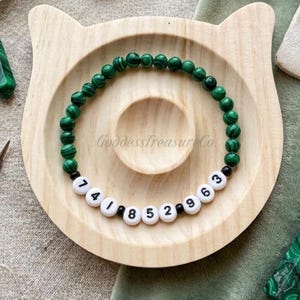 May include: A bracelet featuring green malachite beads and white letter beads with numbers. The bracelet is presented on a wooden cat-shaped tray. The text "GoddessTreasureCo" is visible on the tray.