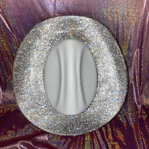 Puede incluir: Un sombrero de ala ancha con una copa central blanca mate y lisa que presenta una marcada hendidura vertical. El ala ancha est&aacute; completamente cubierta de un material de purpurina plateada densa y reflectante.