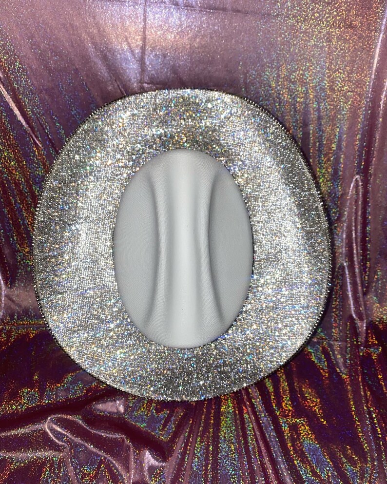 Puede incluir: Un sombrero de ala ancha con una copa central blanca mate y lisa que presenta una marcada hendidura vertical. El ala ancha est&aacute; completamente cubierta de un material de purpurina plateada densa y reflectante.