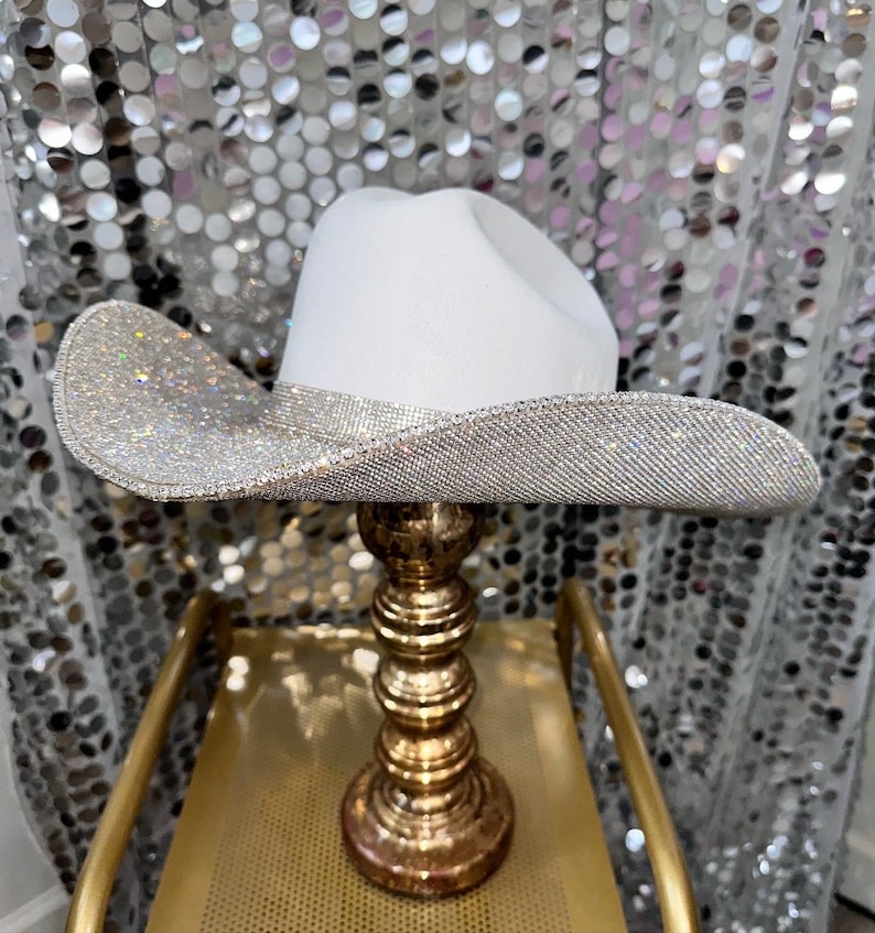 Puede incluir: Un sombrero de vaquero blanco con un ala ancha y curva completamente cubierta de peque&ntilde;os diamantes de imitaci&oacute;n plateados reflectantes que crean un efecto brillante. La copa del sombrero es de color blanco mate liso y el ala tiene un borde plateado.
