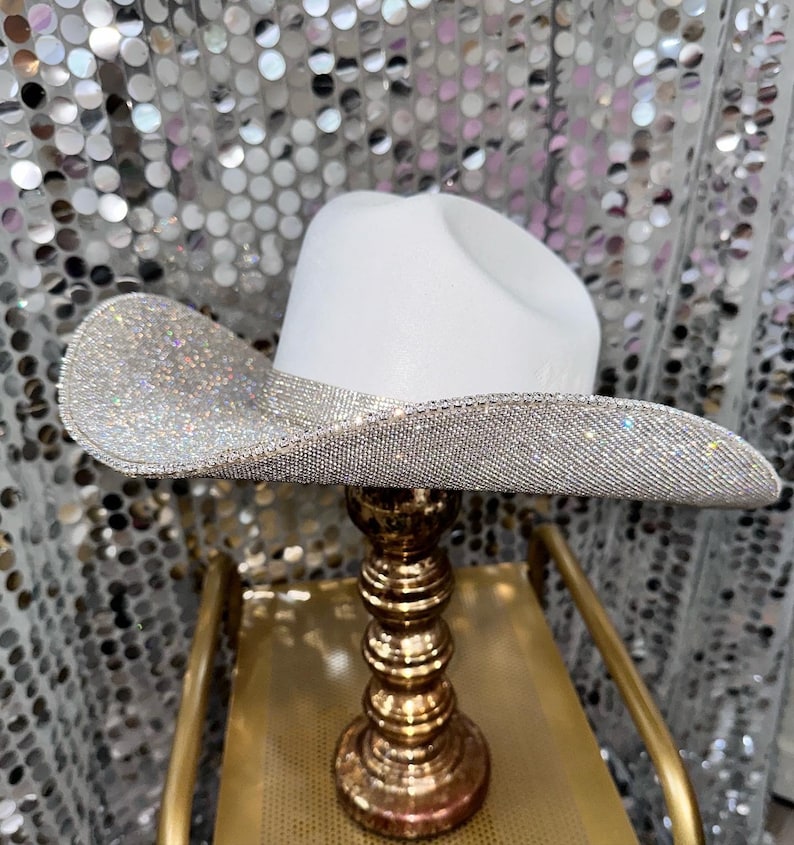 Puede incluir: Un sombrero de vaquero blanco con un ala ancha completamente cubierta de peque&ntilde;os diamantes de imitaci&oacute;n plateados reflectantes que crean un efecto brillante. La copa del sombrero es blanca mate, lisa y tiene un dise&ntilde;o tradicional.