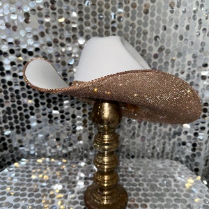 May include: A white cowboy hat with a gold glitter band. The hat is sitting on a gold stand.