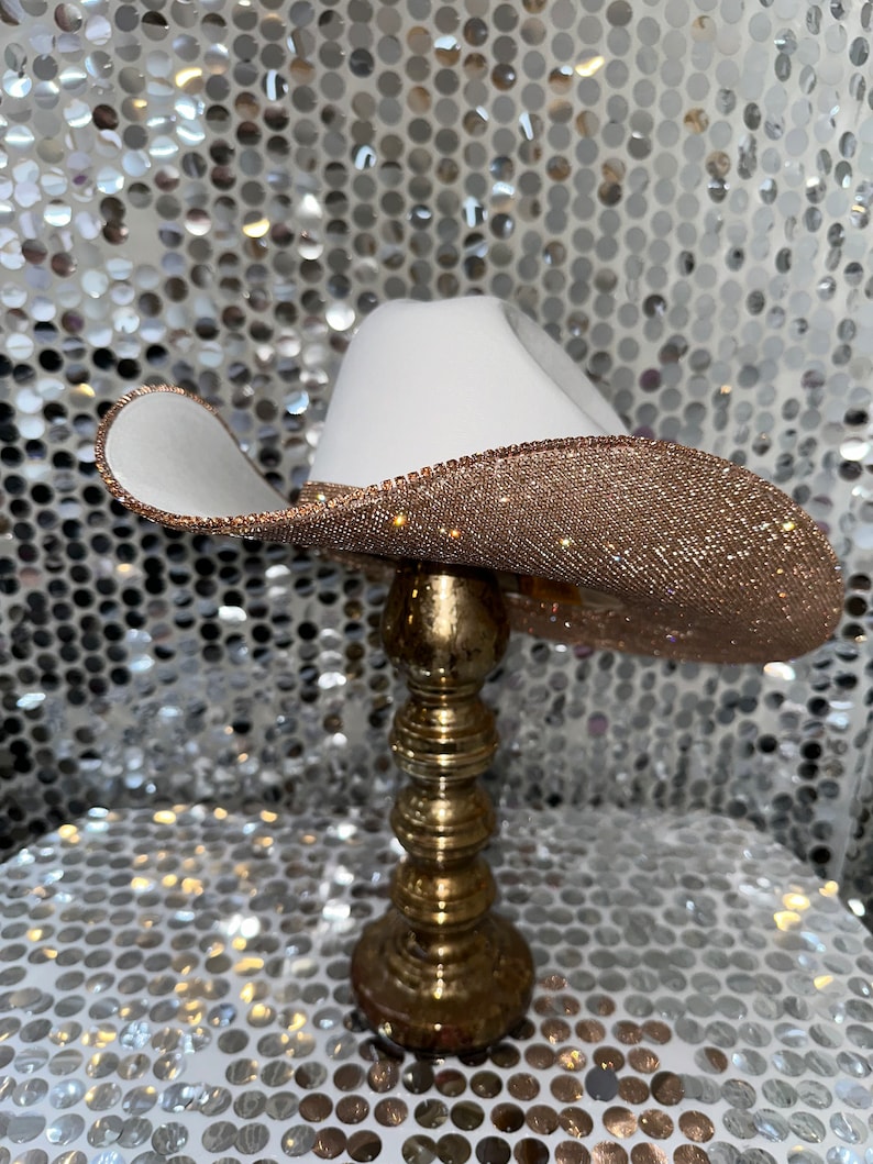 May include: A white cowboy hat with a gold glitter band. The hat is sitting on a gold stand.
