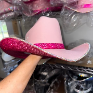 May include: A pink cowboy hat with a wide brim, decorated with a band of sparkling rhinestones. The hat is trimmed with a pink ribbon and has a pink felt crown.