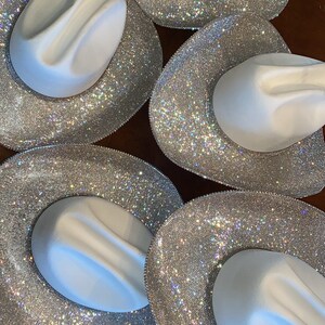Puede incluir: Cinco sombreros de vaquero blancos dispuestos en un patr&oacute;n circular. Cada sombrero presenta una copa blanca lisa y un ala ancha completamente cubierta de peque&ntilde;os diamantes de imitaci&oacute;n plateados que crean un efecto de luz brillante.
