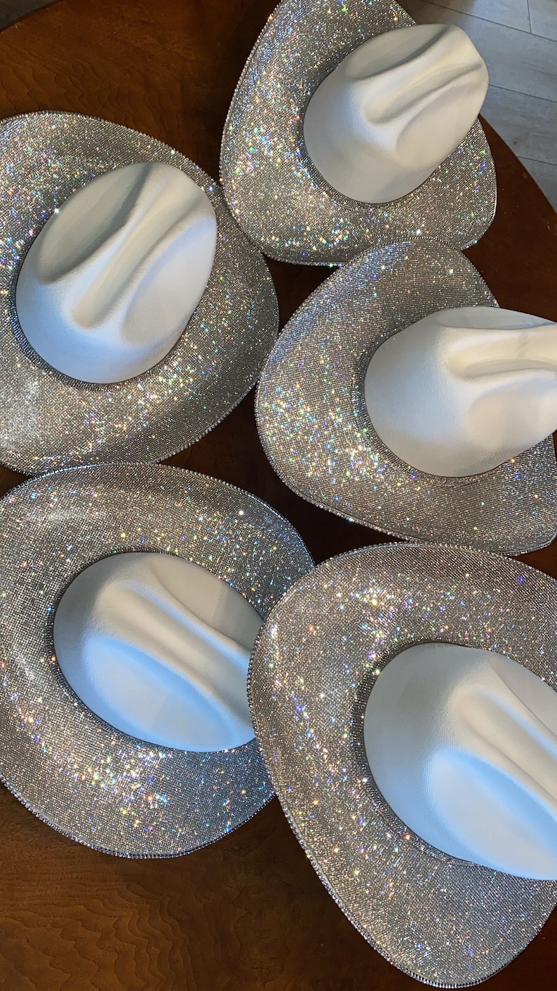 Puede incluir: Cinco sombreros de vaquero blancos dispuestos en un patr&oacute;n circular. Cada sombrero presenta una copa blanca lisa y un ala ancha completamente cubierta de peque&ntilde;os diamantes de imitaci&oacute;n plateados que crean un efecto de luz brillante.