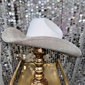 Puede incluir: Un sombrero de vaquero blanco con un ala ancha y curva completamente cubierta de peque&ntilde;os diamantes de imitaci&oacute;n plateados reflectantes. La copa del sombrero es de fieltro blanco mate liso y el borde est&aacute; rematado con una fila de cristales plateados.