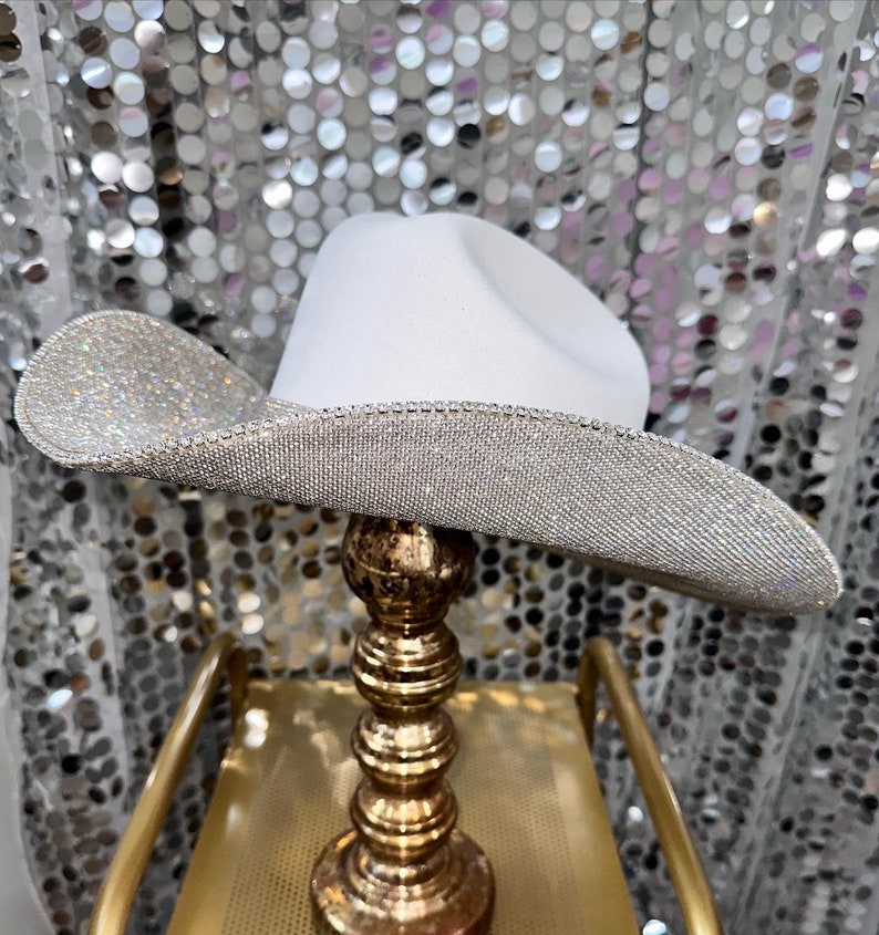 Puede incluir: Un sombrero de vaquero blanco con un ala ancha y curva completamente cubierta de peque&ntilde;os diamantes de imitaci&oacute;n plateados reflectantes. La copa del sombrero es de fieltro blanco mate liso y el borde est&aacute; rematado con una fila de cristales plateados.