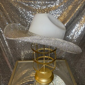 Puede incluir: Un sombrero de vaquero blanco con un ala ancha completamente cubierta de brillantes diamantes de imitaci&oacute;n plateados. La copa del sombrero es lisa y de color blanco mate, lo que contrasta con la textura brillante y reflectante del ala. El sombrero est&aacute; sobre un soporte dorado.
