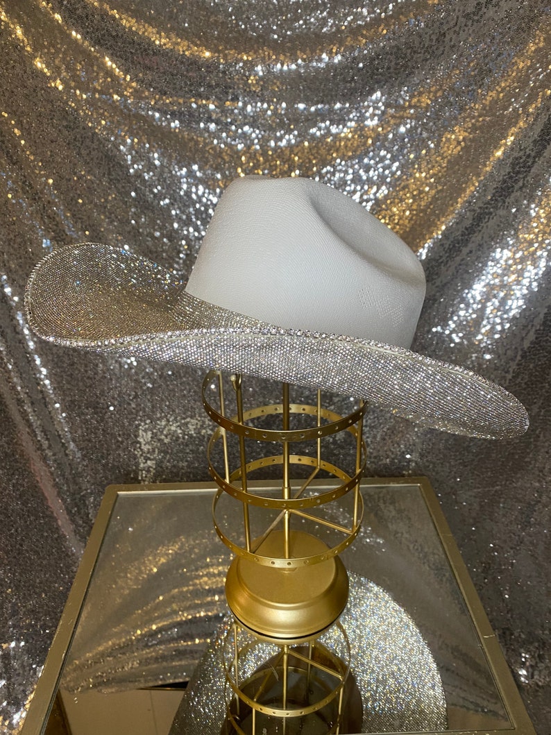Puede incluir: Un sombrero de vaquero blanco con un ala ancha completamente cubierta de brillantes diamantes de imitaci&oacute;n plateados. La copa del sombrero es lisa y de color blanco mate, lo que contrasta con la textura brillante y reflectante del ala. El sombrero est&aacute; sobre un soporte dorado.