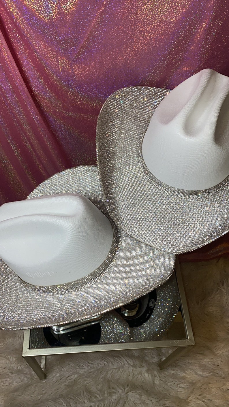 Puede incluir: Dos sombreros de vaquero blancos con un acabado brillante en la copa y un ala completamente cubierta de densos y brillantes diamantes de imitaci&oacute;n plateados que reflejan la luz, creando un efecto centelleante en la superficie.