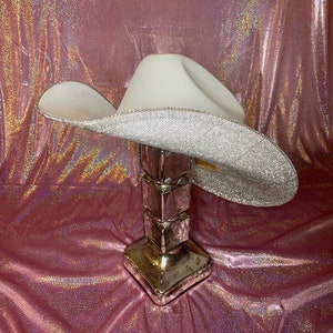 Puede incluir: Un sombrero de vaquero blanco con un ala ancha completamente cubierta de brillantes diamantes de imitaci&oacute;n plateados. La copa del sombrero es lisa y de color blanco mate, mientras que la parte inferior del ala muestra una textura densa y brillante.