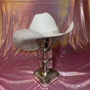 Puede incluir: Un sombrero de vaquero blanco con un ala ancha cubierta de brillantes diamantes de imitaci&oacute;n plateados. El sombrero incluye una banda de pedrer&iacute;a a juego que rodea la base de la copa, con los bordes del ala tambi&eacute;n detallados con pedrer&iacute;a.