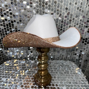 May include: A white cowboy hat with a gold rhinestone trim. The hat is sitting on a gold stand.