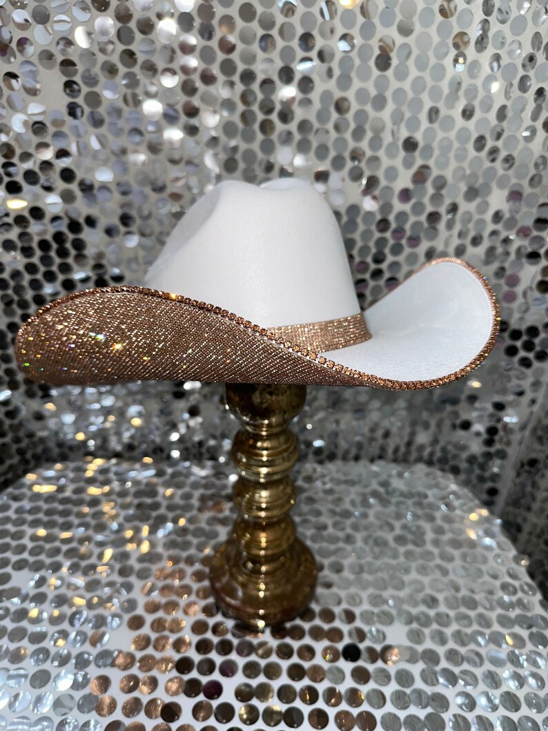 May include: A white cowboy hat with a gold rhinestone trim. The hat is sitting on a gold stand.