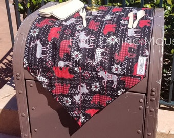 May include: A red and black plaid table runner with white snowflakes and reindeer designs. The runner is on top of a brown metal trash can. A white candle, a small white box, and two wooden utensils are on the table runner.