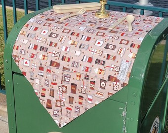 May include: A green metal trash can with a brown and white coffee cup patterned tablecloth on top. The tablecloth has a white candle and a wooden spoon and fork on it. The trash can has a sticker that says "Please Help Keep Our City Clean" with a cartoon image of a building.
