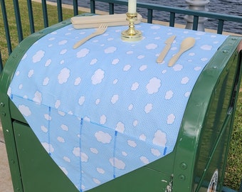 May include: A green trash can with a blue and white tablecloth on top. The tablecloth has a cloud pattern. The trash can has a sticker that says "Please Help Keep Our City Clean".